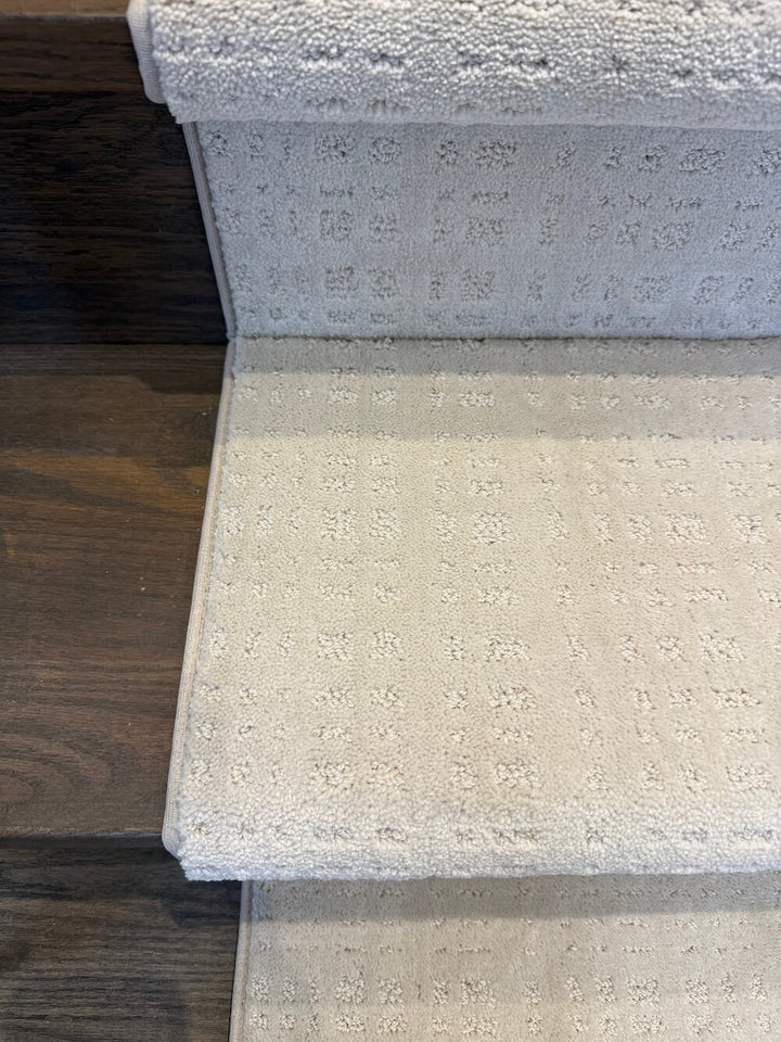 Close-up of a light beige or off-white carpeted staircase step with a textured, embossed floral or geometric pattern, shown next to a dark wood floor.