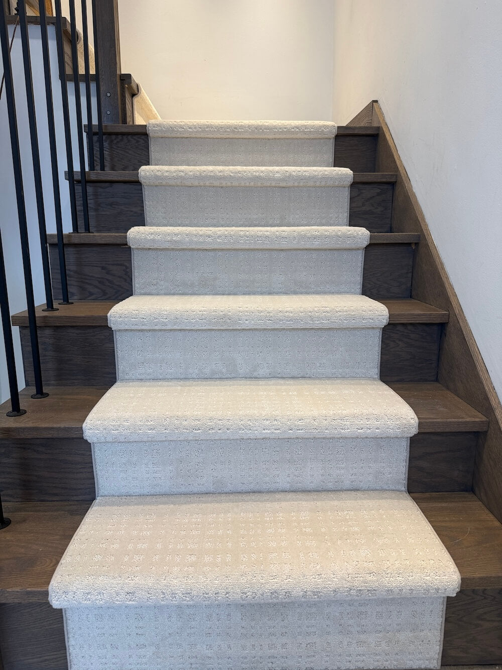 A set of indoor staircase steps covered in light beige textured carpet, with dark wood treads and a black metal railing on the left against a white wall.