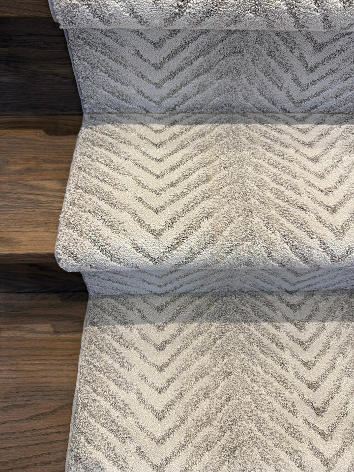 A close-up of carpeted stairs with a textured chevron pattern in light gray and off-white, showing the transition from dark wood flooring at the left edge.