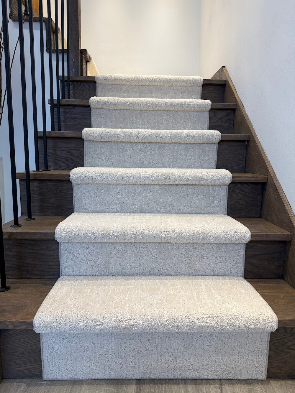 A staircase with light beige textured carpeted steps and dark wood risers, framed by a black metal railing against a white wall.