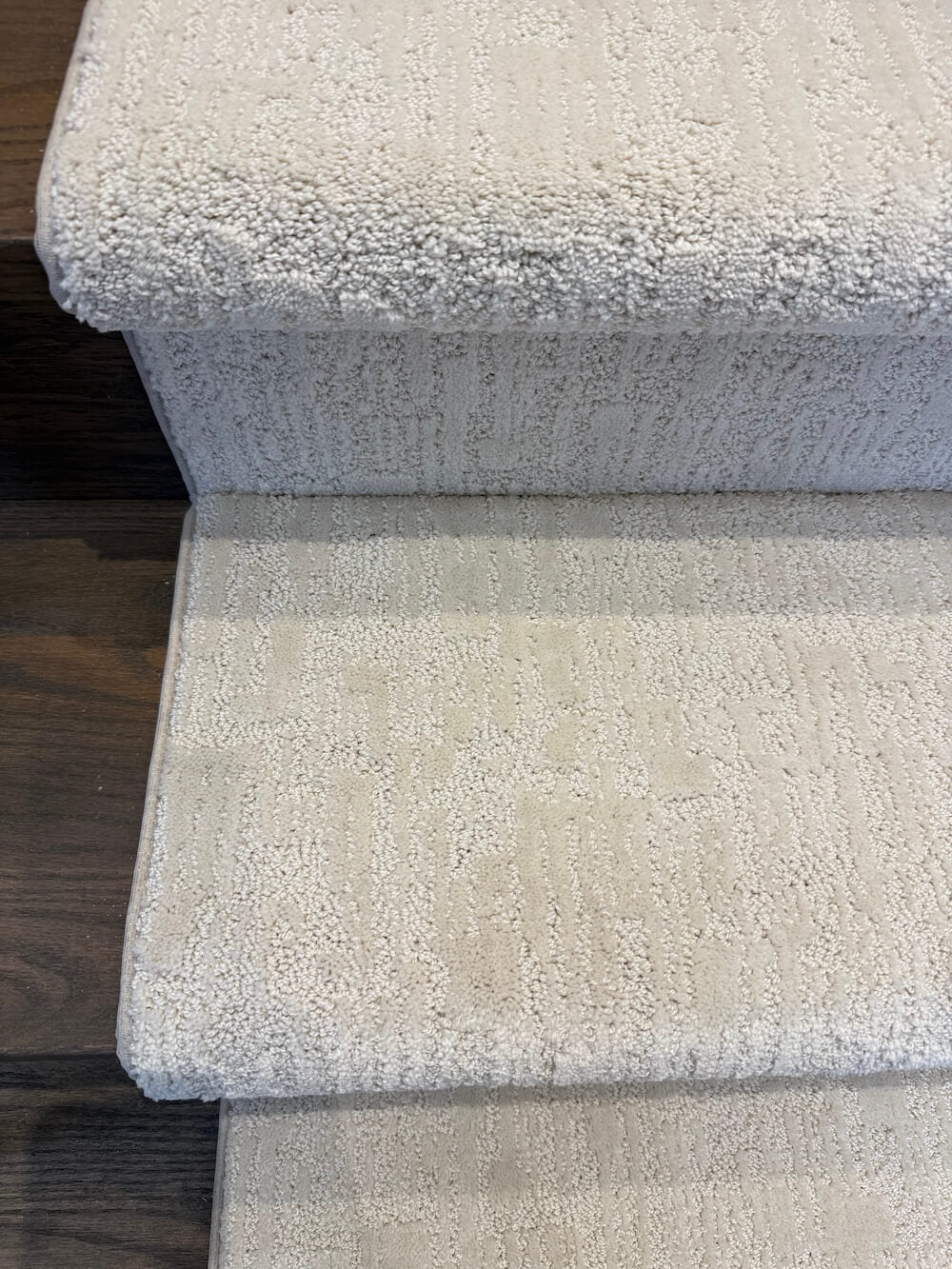 Close-up of carpeted stairs in a light beige or off-white textured fabric, with dark wood flooring visible at the edges.