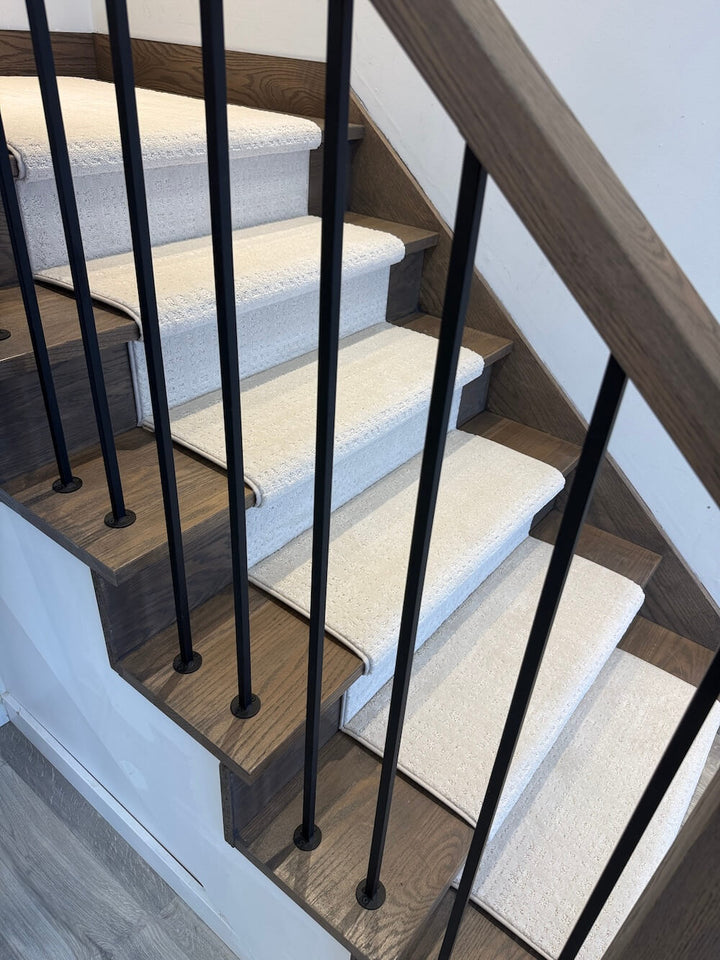 A close-up view of an indoor staircase with light beige carpeted steps, medium brown wood risers, black metal balusters, and a wooden handrail against a white wall.