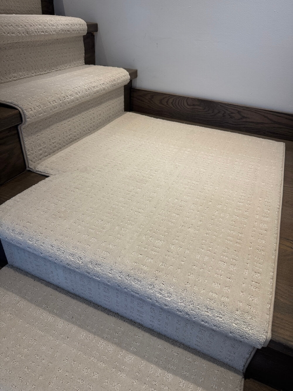 A light beige textured carpet runner or stair carpet shown installed on a set of indoor stairs, with a matching flat section laid out on the floor in front.